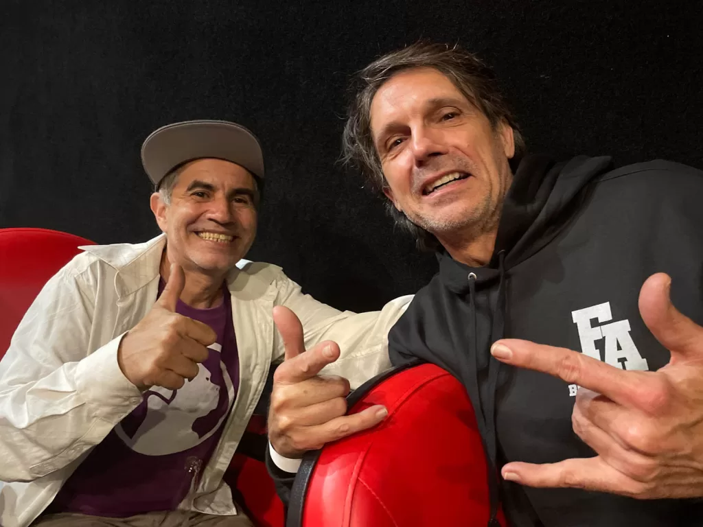 Foto de dois homens sorrindo e fazendo sinal de positivo na câmera, um usando boné e o outro com camiseta esportiva, em um ambiente interno.