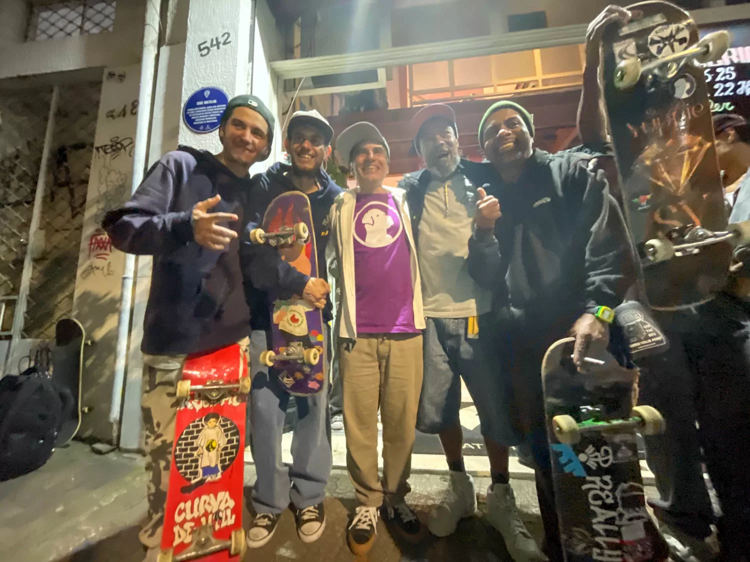 Grupo de skatistas com skateboards em uma rua à noite, comemorando após uma sessão de skate, vestindo roupas casuais e sorrindo para a câmera.