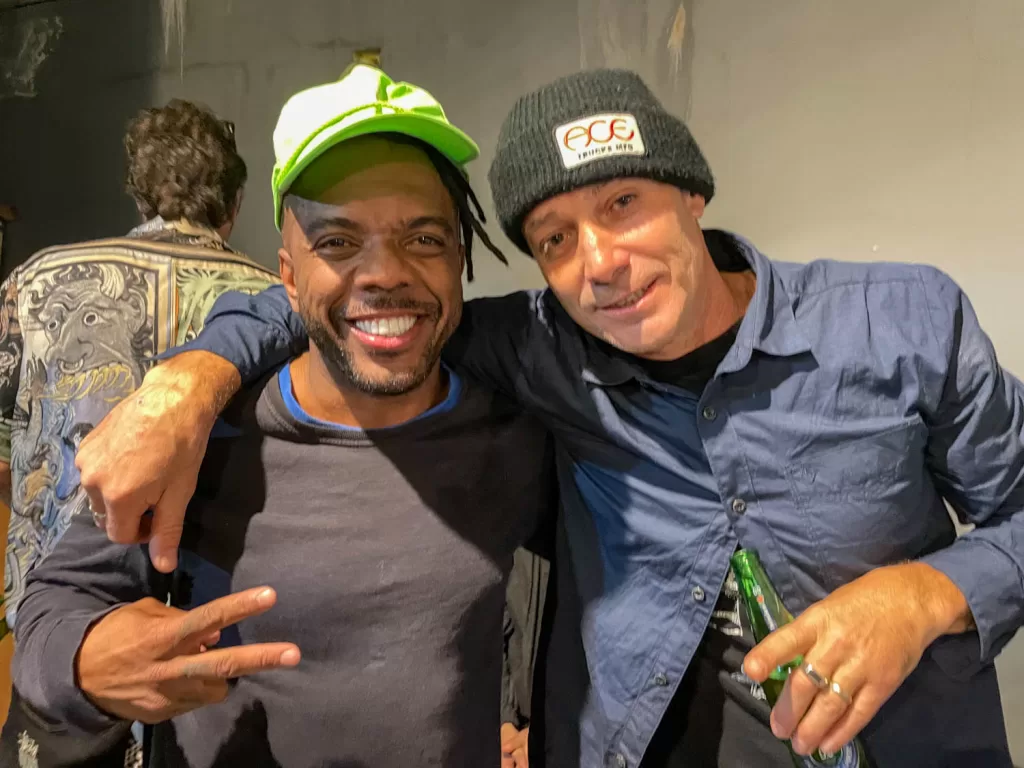 Duas pessoas sorrindo, brincando e comemorando juntos em um ambiente social, com um fundo artístico e descontraído.