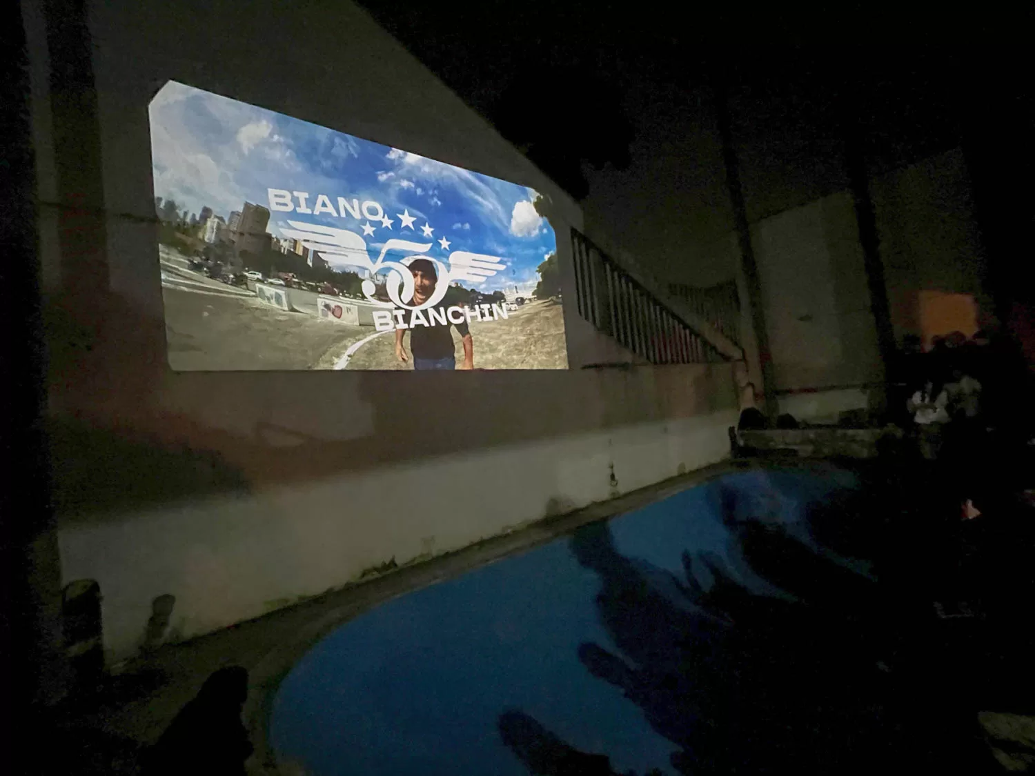 Vídeo de skate do Biano Bianchin projetado em uma parede de fundo azul escuro, com uma pessoa na frente, em uma área de eventos ou reunião.
