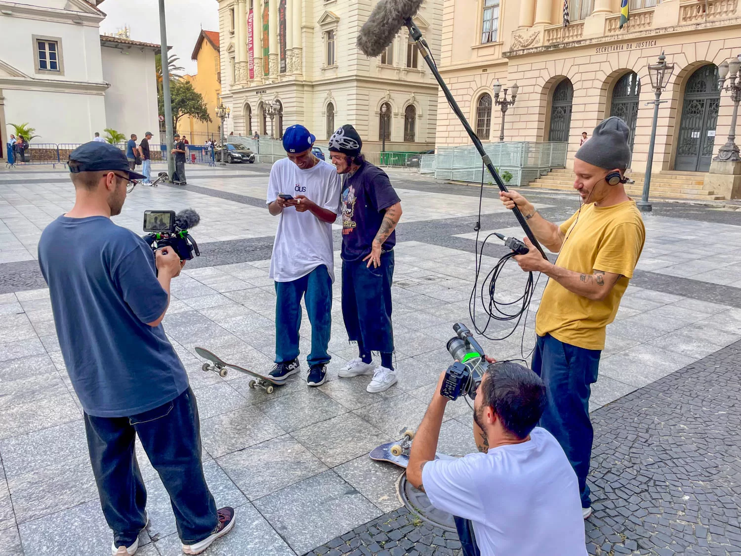 Equipe de produção de vídeo grava cena ao ar livre com um skate em uma praça histórica, enquanto duas pessoas observam e utilizam celulares, cenário urbano com edifícios antigos ao fundo.